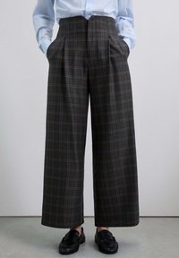 Pantalones de cuadros marrones con piernas anchas, pliegues en la parte delantera y bolsillos laterales, hechos de un tejido texturizado, combinados con mocasines negros.