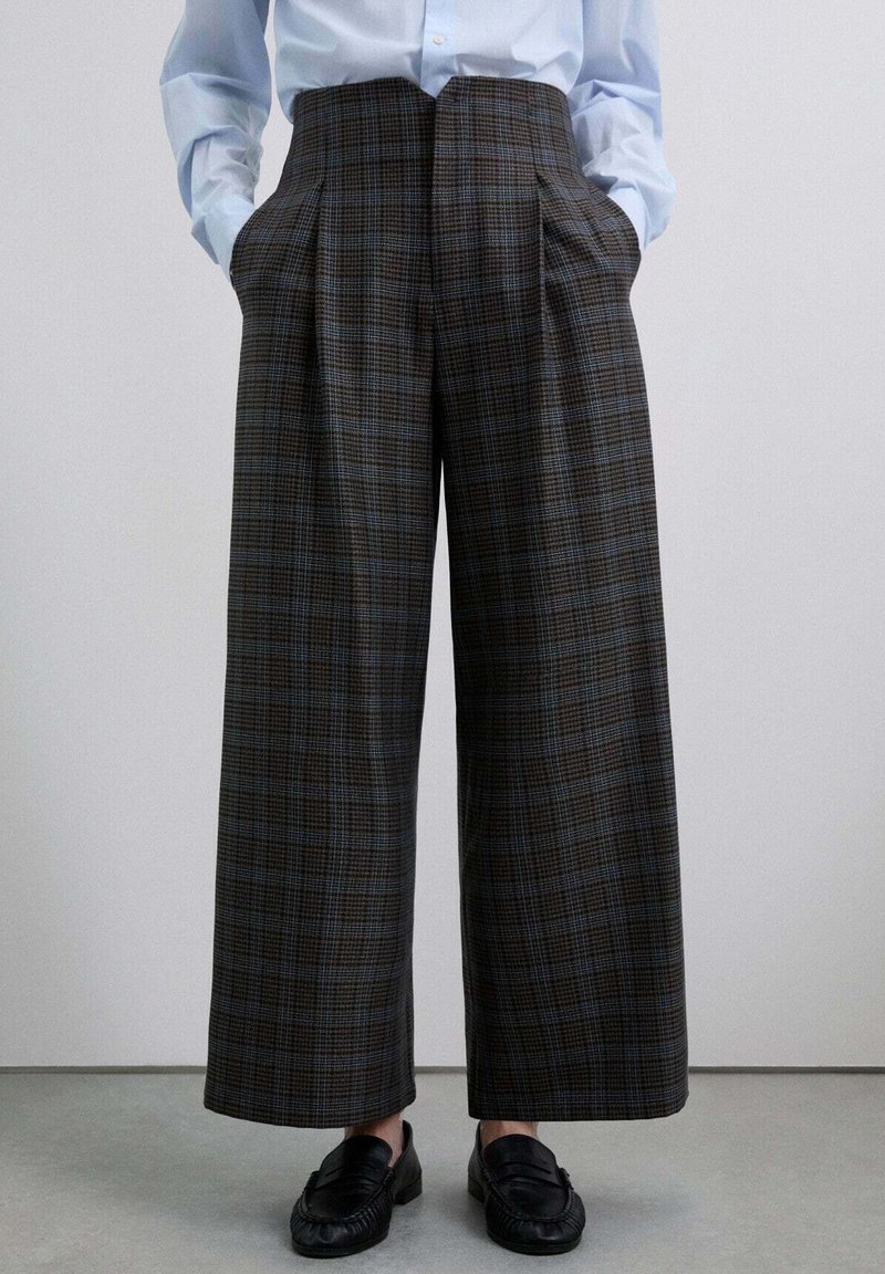 Pantalones de cuadros marrones con piernas anchas, pliegues en la parte delantera y bolsillos laterales, hechos de un tejido texturizado, combinados con mocasines negros.
