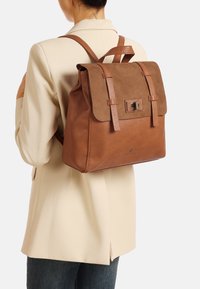 Zaino in pelle marrone con patta in suede, dotato di doppie cinghie, hardware in metallo e finitura texturizzata, esposto su uno sfondo neutro.