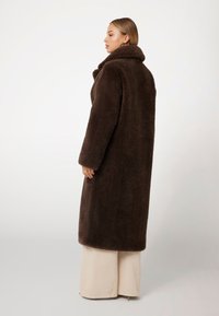 Abrigo de piel sintética marrón con un cuello ancho, mangas largas y corte recto, combinado con pantalones de pierna ancha beige. Material suave y texturizado.