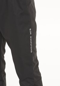 Pantalons de sport noirs avec une texture lisse, arborant les mots "ENDURANCE" et "XCS" en blanc sur la jambe inférieure.