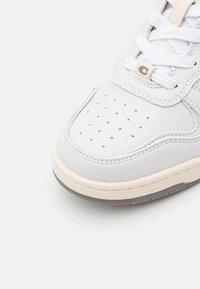 Vita lädersneakers med perforerad tå, matchande vita snören och en kontrasterande beige och grå sula. Subtil logotypdetalj är inkluderad.