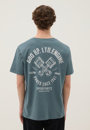 Jong persoon draagt een blauw T-shirt met gekruiste motorzuigers en tekst over pk, vermogen en snelheidsonderdelen op de achterkant.