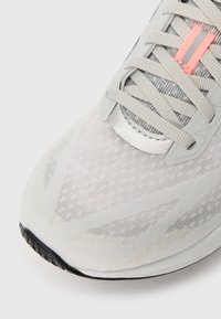 Gros plan sur une chaussure de sport blanche avec des lacets gris clair, une boucle de lacet argentée et rose, et une semelle texturée avec un bout noir.