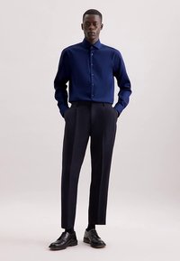 Marineblauw overhemd met knopen en een gladde textuur, gecombineerd met hooggetailleerde, donkergrijze op maat gemaakte broek. De benen hebben verticale strepen. Zwarte schoenen.