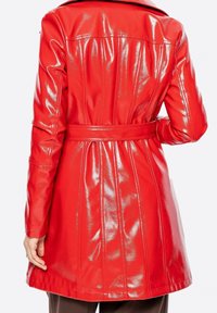 Femme portant un trench-coat en similicuir rouge brillant, vue de dos avec une ceinture cintrant la taille et des manches longues.