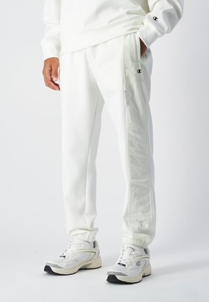 Pantalones de chándal blancos con puños elásticos, detalles en los paneles laterales y un logotipo en la parte frontal. Combinados con zapatillas blancas ligeras que tienen detalles en negro.