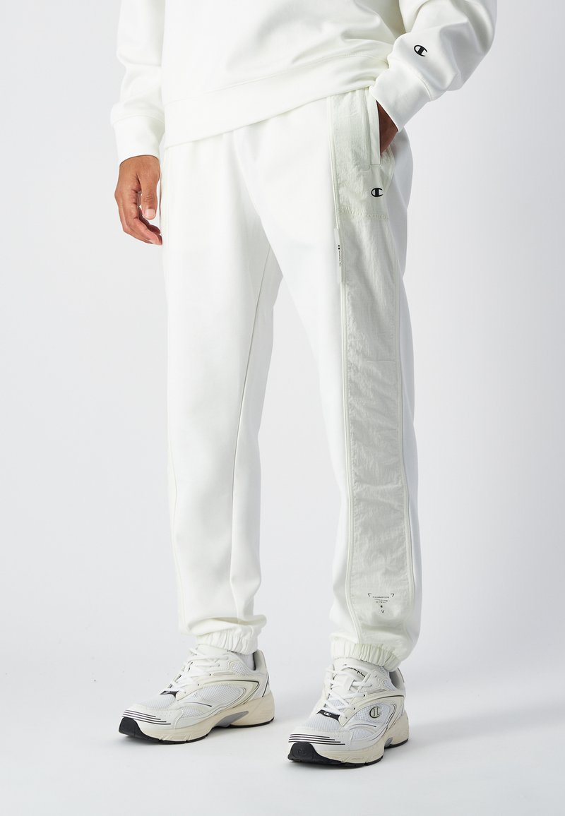 Pantalons de survêtement blancs avec des poignets élastiques, des détails de panneaux latéraux et un logo à l'avant. Assortis à des baskets légères blanches avec des accents noirs.