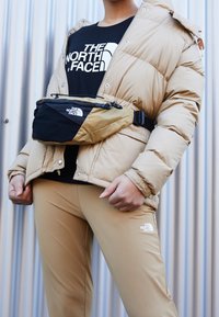 Osoba v béžové péřové bundě, béžových kalhotách, černém tričku The North Face a béžovo-černé ledvince The North Face před vlnitou kovovou zdí.