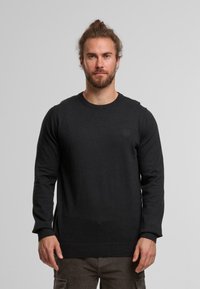 Suéter de punto negro con cuello redondo y mangas largas. Presenta puños y dobladillo acanalados, con un sutil logotipo en el lado izquierdo del pecho. Textura suave.