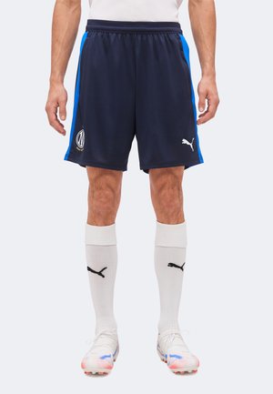 Muž, ktorý nosí tmavomodré športové šortky s modrými bočnými pruhmi, biele logo Puma, biele podkolienky a biele kopačky s modrými a červenými detailmi.
