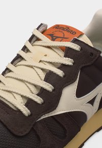 Κοντινό πλάνο καφέ και κρεμ αθλητικού παπουτσιού Mizuno Runbird με κορδόνια κρεμ, υλικά από δέρμα και δίχτυ, και πορτοκαλί ετικέτα με λογότυπο στη γλώσσα.