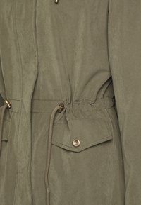 Chaqueta verde oliva con acabado texturizado, que cuenta con cintura con cordón, bolsillo con broche y detalles en herrajes dorados.