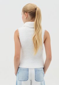 Suéter blanco sin mangas de cuello alto, hecho de una tela difusa con una textura de canalé. El dobladillo está ligeramente deshilachado. Combinado con jeans de mezclilla claros.