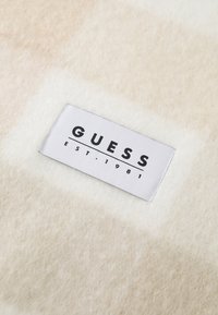 Fleece-stof i beige og kremfarver med et hvidt label med "GUESS EST. 1981" i sort tekst. Blød tekstur og minimalistisk design.