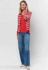 Femme souriante aux cheveux blonds portant un cardigan à rayures rouges, un haut rouge, un jean bleu à jambes larges et des chaussures plates beiges, debout devant un fond blanc.