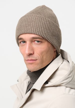 Gorro de punto acanalado beige con una parte superior ligeramente puntiaguda, llevado sobre un cuello de tortuga oscuro, combinado con una chaqueta de color claro.