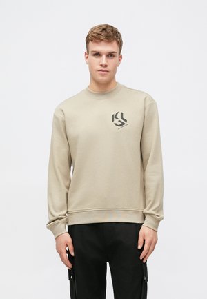 Sweatshirt beige à manches longues, col rond et logo graphique noir sur la poitrine gauche. Associé à un pantalon noir avec poches cargo.