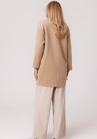 Beżowa tunika z długim rękawem o prostym kroju i subtelnym przeszyciu. Połączona z luźnymi, jasnobeżowymi spodniami. Miękka, gładka tekstura tkaniny.