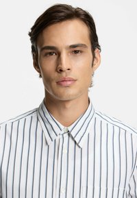 Weißes Hemd mit Knopfleiste, vertikalen blauen Streifen, spitzem Kragen und Tasche auf der linken Brustseite. Glatter Baumwollstoff mit einem knackigen Aussehen.