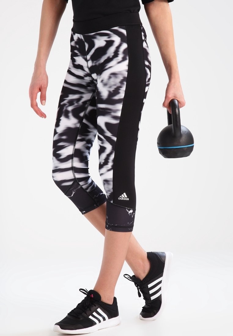 Personne portant des leggings noirs et blancs à motifs et des baskets Adidas noires, tenant un kettlebell noir avec une bande bleue dans une main.