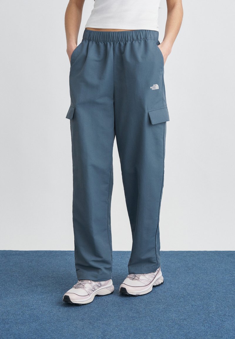 Άτομο που φορά μπλε παντελόνι καμπαρτίνας με ελαστική μέση και λευκά αθλητικά παπούτσια, στέκεται σε μπλε χαλί με τα χέρια στις τσέπες.