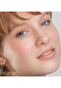 Gros plan sur un visage à la peau claire, avec des taches de rousseur légères et un maquillage doux et naturel. Elle a les cheveux brun clair et un sourire subtil. Boucles d'oreilles en or.