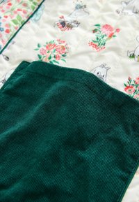 Groen corduroy stof op een gewatteerd witte ondergrond met bloemen- en cartoonfiguurafdrukken in roze, groen en rode kleuren.
