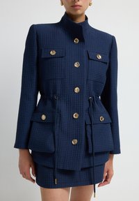 Granatowa kurtka o strukturze z kołnierzem stawianym, złotymi guzikami, czterema kieszeniami z klapkami i sznurkiem w pasie.