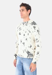 Maglione girocollo color crema con motivo marmorizzato nero, texture lavorata a maglia e polsini a costine. Abbinato a jeans di denim azzurro chiaro.