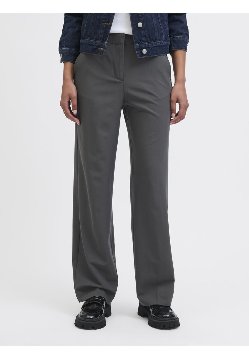 Femme portant un pantalon large gris foncé associé à des chaussures noires épaisses et une veste en jean sur une chemise blanche.