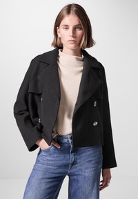 Svart kort dubbelknäppt jacka med stora slag, silverknappar, bärs över en beige polotröja, parat med blå denimjeans.