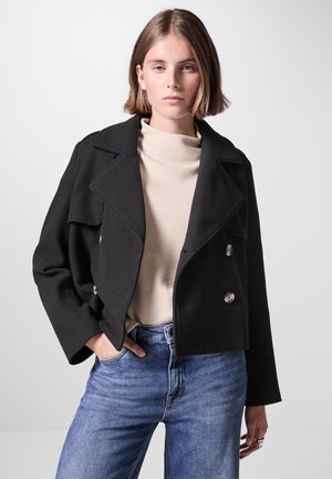 Schwarze, verkürzte, doppelt geknöpfte Jacke mit großen Revers, silbernen Knöpfen, getragen über einem beigen Rollkragenpullover, kombiniert mit blauen Jeans.