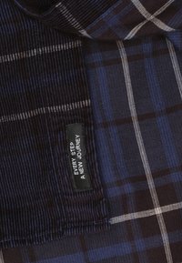 Houten stof in blauw en donkerbruin, met witte lijnen. Bevat een zwart label met de tekst "IEDERE STAP EEN NIEUWE REIS." Textuurkatoen.