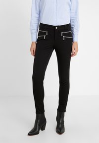 Pantalones negros ajustados con dos bolsillos frontales con cremallera, elaborados con una tela suave. El modelo lleva botas de tobillo negras, que añaden un toque elegante.