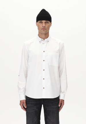 White long-sleeve button-up shirt with a single chest pocket, bronze buttons, and a smooth texture, paired with dark jeans and a black beanie.
