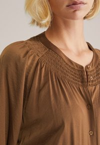 Brun knapplång blus i mjukt tyg. Har en smockad design vid halsringningen och rund krage, med en avslappnad, flödande passform.