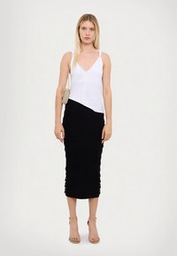 Moteris stovi vilkėdama baltą be rankovių marškinėlį, juodą midi sijoną, smėlio spalvos aukštakulnius sandalus ir laiko mažą aukso grandinėlės rankinę.