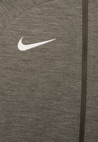 Graues Sportoberteil aus strukturiertem Stoff. Verfügt über ein weißes Nike-Logo und einen vertikalen Reißverschluss auf der Vorderseite.