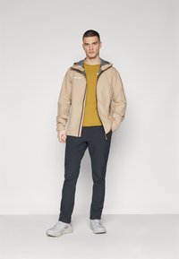 Beige lättjacka med grå huva, svarta dragkedjor, bärs över en senapsgul skjorta och tillsammans med mörka byxor och grå skor. Slät textur.