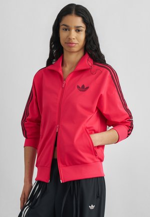 Frau trägt eine rote Adidas Trainingsjacke mit schwarzen Streifen und schwarze Adidas Hose mit weißen Streifen, steht mit einer Hand in der Tasche.