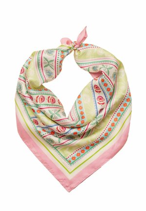 Kobieta w żółtym t-shircie z nadrukiem graficznym, pastelowych, szerokich spodniach w paski, niebieskim szydełkowanym kapeluszu, pomarańczowym naszyjniku z koralików i żółtych sandałach, stojąca na dywanie.
