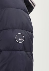 Granatowa kurtka puchowa z okrągłym czarnym logo z napisem "edc", pionowym przeszyciem oraz gładką, błyszczącą teksturą materiału.