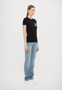 T-shirt nera a maniche corte con stampa grafica, abbinata a jeans azzurri e sneakers bianche. Il modello si trova di fronte a uno sfondo neutro.