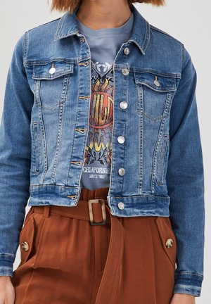 Veste en jean avec boutons argentés sur un t-shirt bleu graphique, associée à un pantalon marron taille haute avec ceinture en tissu et poches boutonnées.