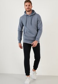 Sweatshirt à capuche gris avec une poche frontale, des cordons de serrage et des poignets côtelés, associé à un jogging noir et des baskets blanches sur un fond uni.