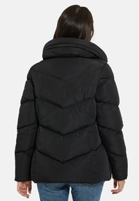 Svart puffjacka med hög krage, sicksack-sömnad och en texturerad yta, bärs över blå jeans.