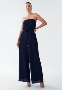 Temno moderen skok z brez naramnicami in plisiranim zgornjim delom, pasom z vrvico, širokimi hlačnicami, lahkotno tkanino in subtilnim sijajem.