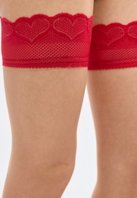 Κόκκινες δαντελένιες κάλτσες μέχρι το μηρό με κορυφές διακοσμημένες με σχέδια καρδιών, φορεμένες σε γυμνά πόδια πάνω σε απλό φόντο.