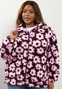 Polarowa kurtka z wysokim kołnierzem, w fioletowej podstawie i dużymi różowymi wzorami kwiatowymi. Zapięcie na zamek z przodu oraz pluszowa tekstura.